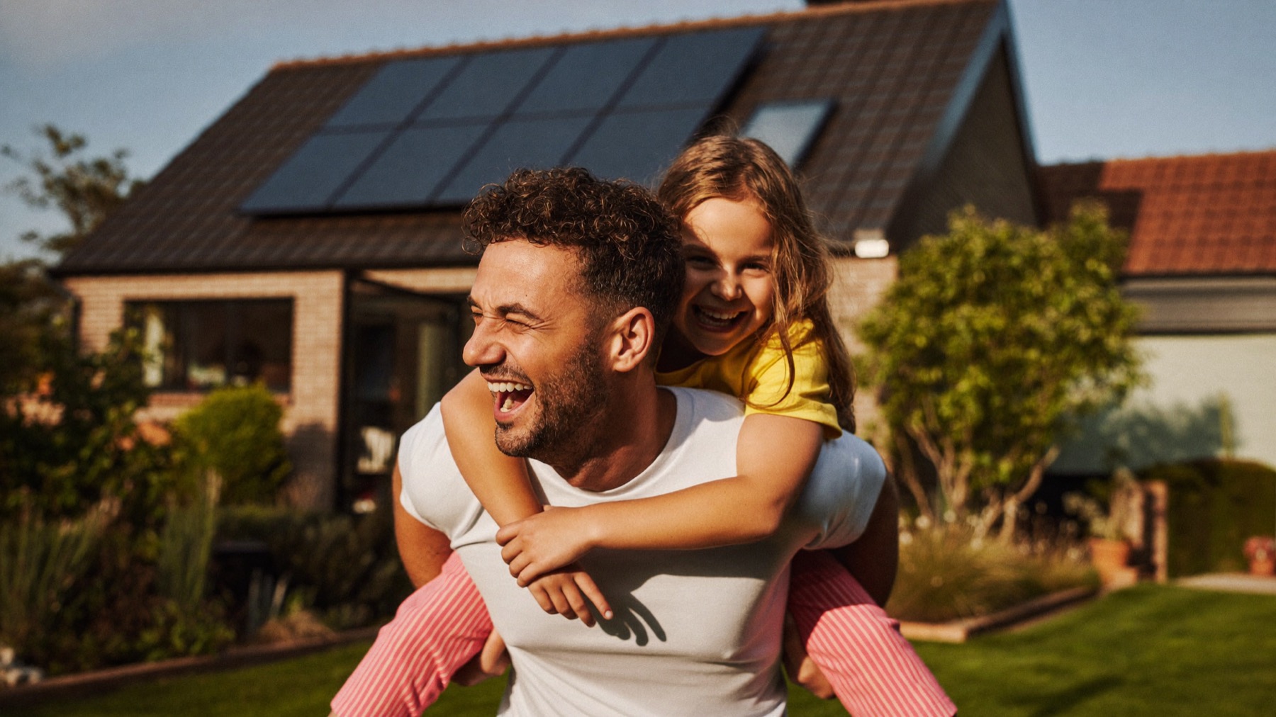Vader en dochter lachend in de tuin voor hun woning met zonnepanelen op het dak